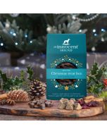 The Innocent Hound Christmas treat box for dogs, displayed on a wooden board with an assortment of treats in the foreground.