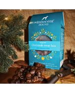 The Innocent Hound Christmas treat box for dogs, displayed on a wooden board with an assortment of treats in the foreground.