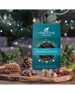 The Innocent Hound Christmas treat box for dogs, displayed on a wooden board with an assortment of treats in the foreground.